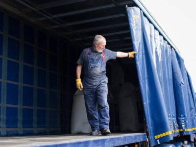 A delivery driver standing on the back of an articulated lorry delivering sustainable fertiliser to a farm in Northumberland in the northeast of England. He is pulling back the lorry curtain to close the trailer.
